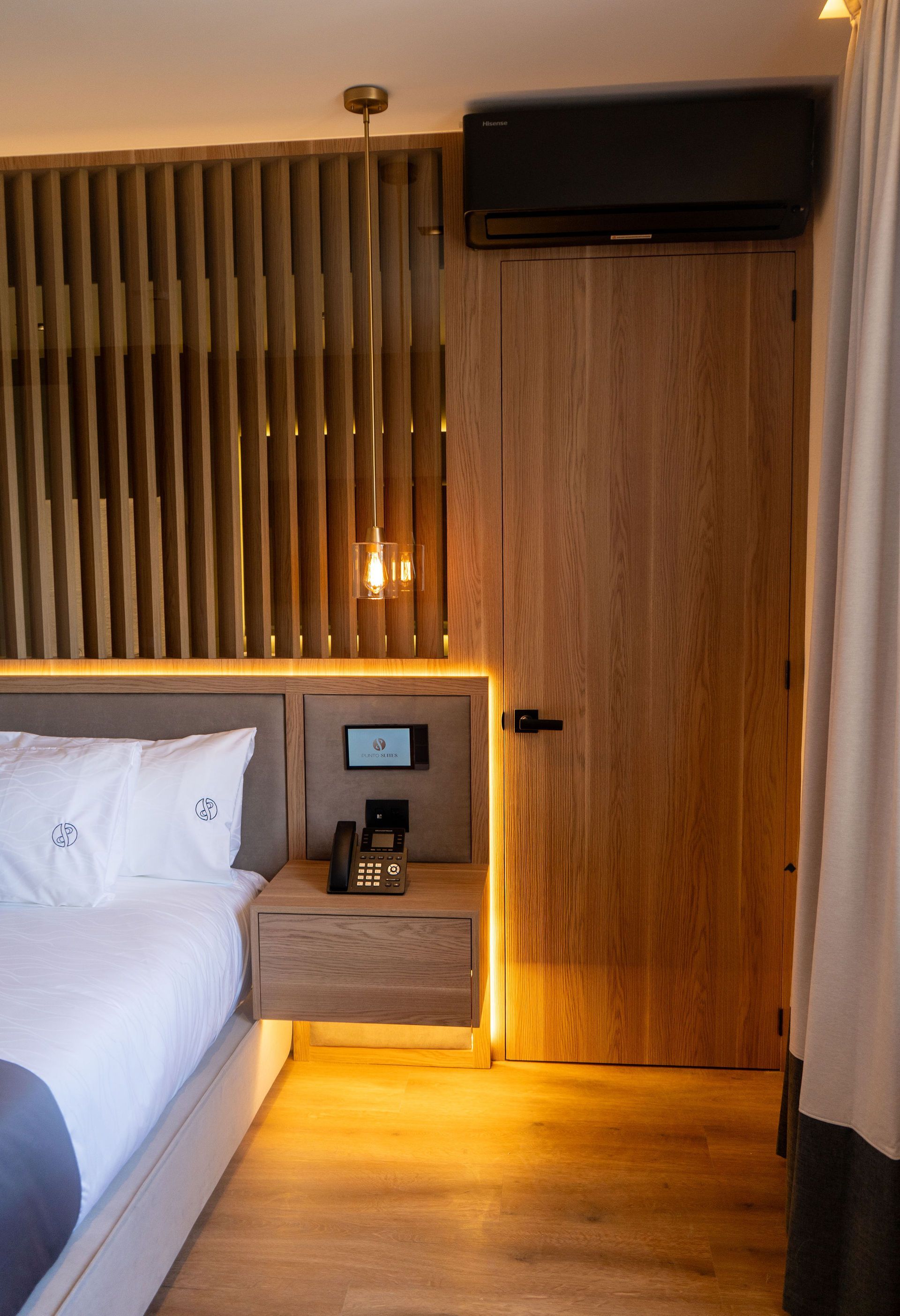 Un rincón de dormitorio moderno con paredes revestidas de madera, una mesita de noche montada en la pared, una lámpara colgante y una puerta de madera.