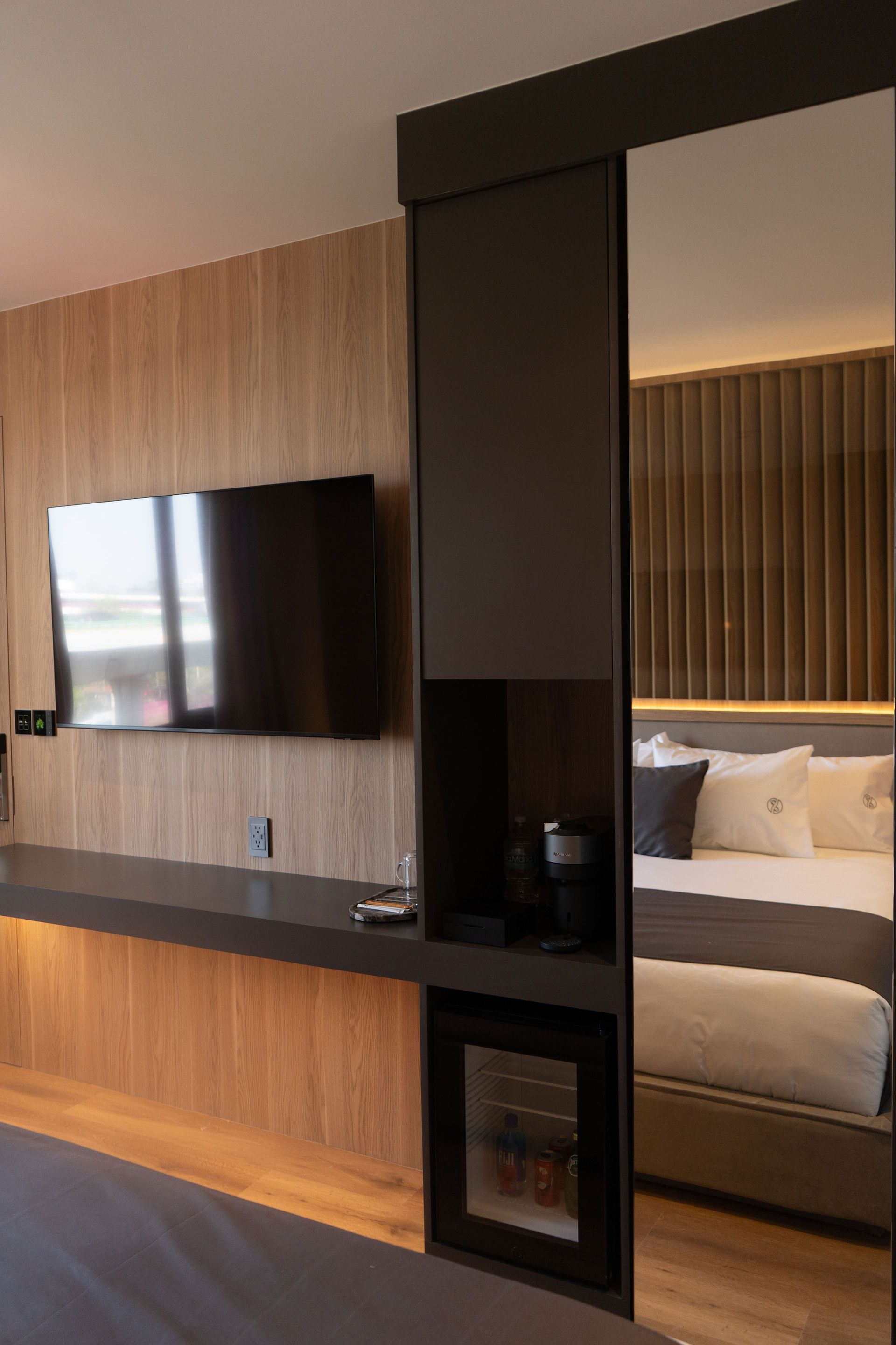 Se refleja una habitación de hotel moderna con un televisor montado en la pared, una estantería, una cafetera integrada y un minibar cerca de la cama.