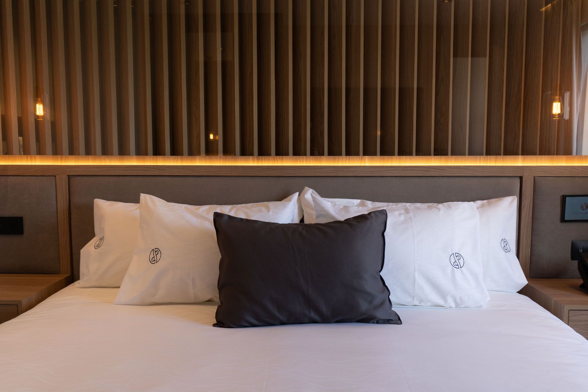 Una cama con almohadas blancas y un cojín central gris oscuro, sobre un cabecero de madera con iluminación cálida empotrada.