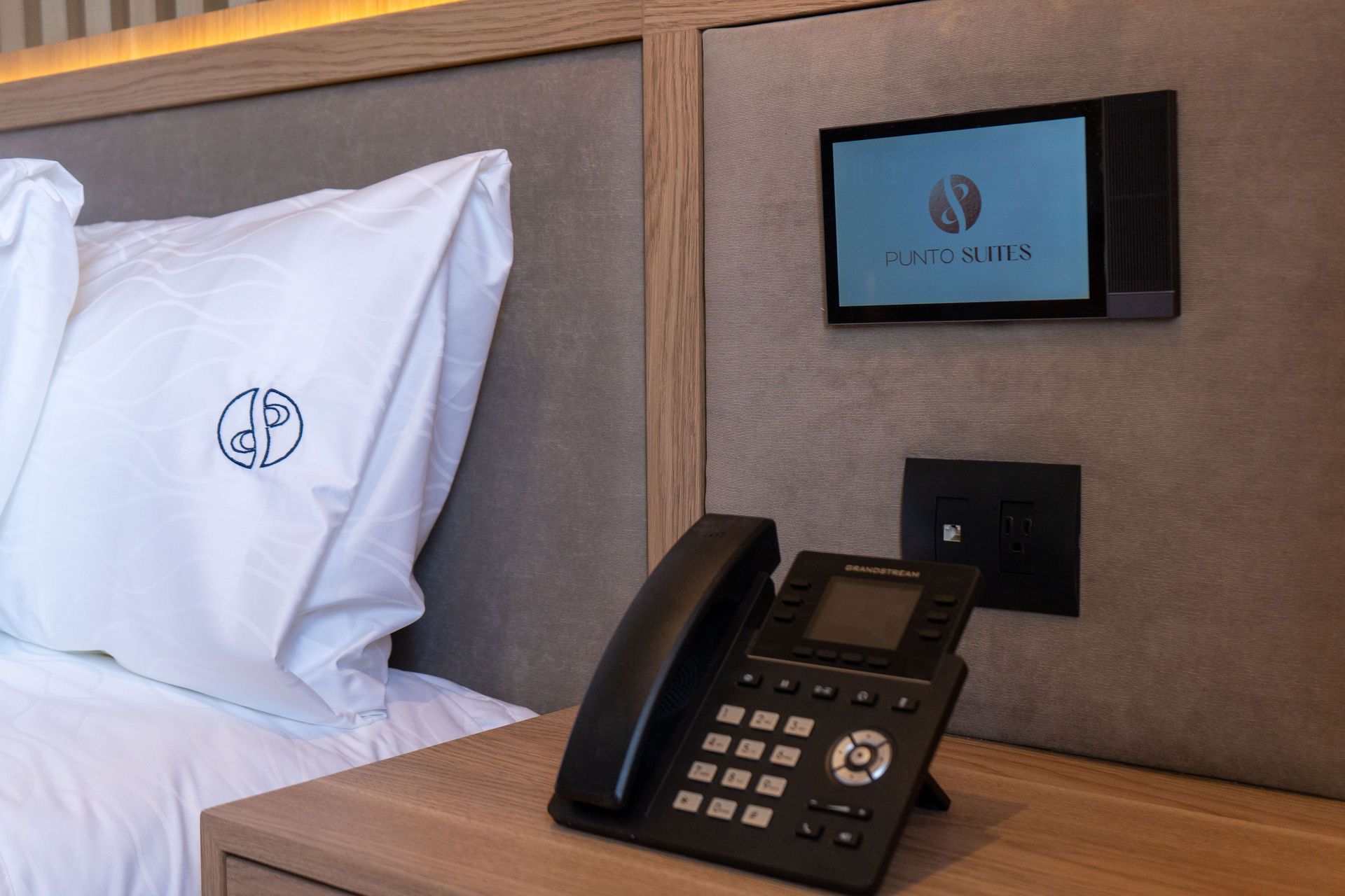 Una mesita de noche de hotel con teléfono fijo, un panel de control táctil montado en la pared y una almohada con un logotipo.