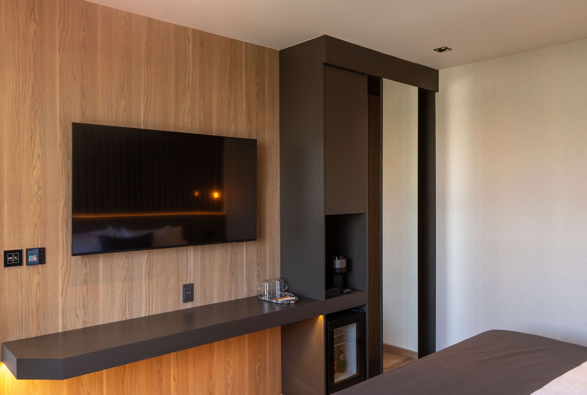 La pared de una habitación de hotel cuenta con un televisor de pantalla plana montado sobre paneles de madera, un escritorio oscuro y una pequeña zona de bar integrada con nevera.