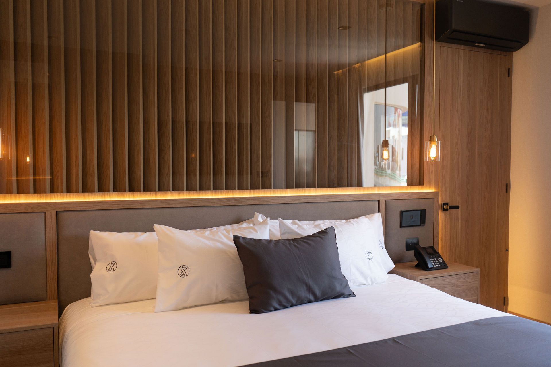 Una habitación de hotel moderna con una cama bien hecha, almohadas blancas, un cojín decorativo oscuro y paneles de madera de color claro.