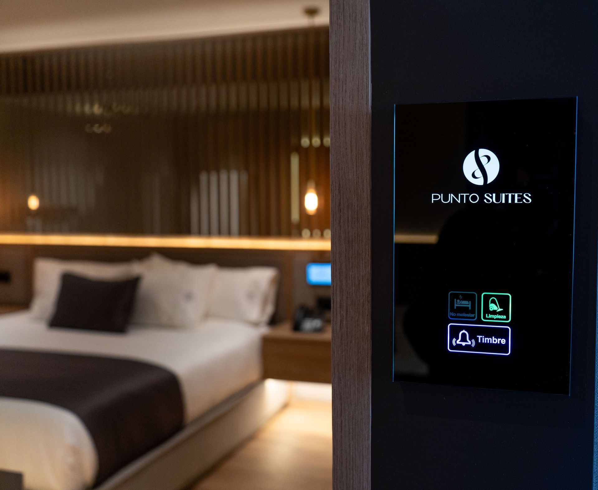 En primer plano se ve el panel de control de una habitación de hotel con botones de servicio iluminados, y detrás, una habitación moderna y difuminada.