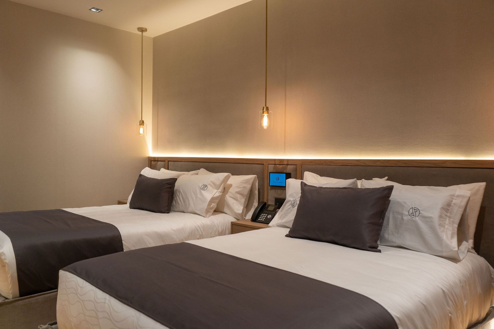 Una habitación de hotel moderna con dos camas tamaño queen, alfombras de color gris oscuro, decoración neutra y lámparas colgantes sobre el cabecero.