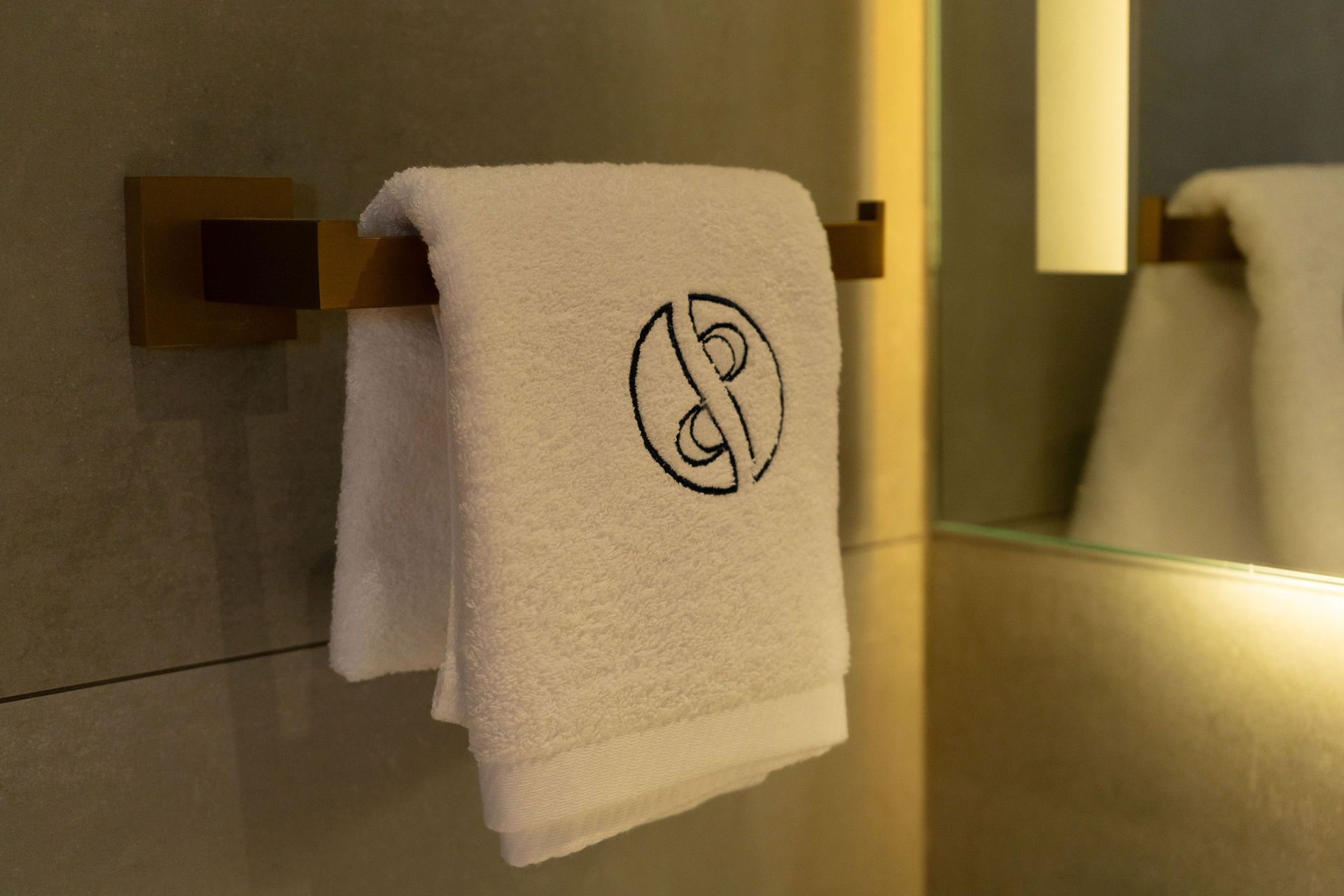 Una toalla blanca con un logotipo circular negro cuelga de un toallero de color bronce en un baño moderno.