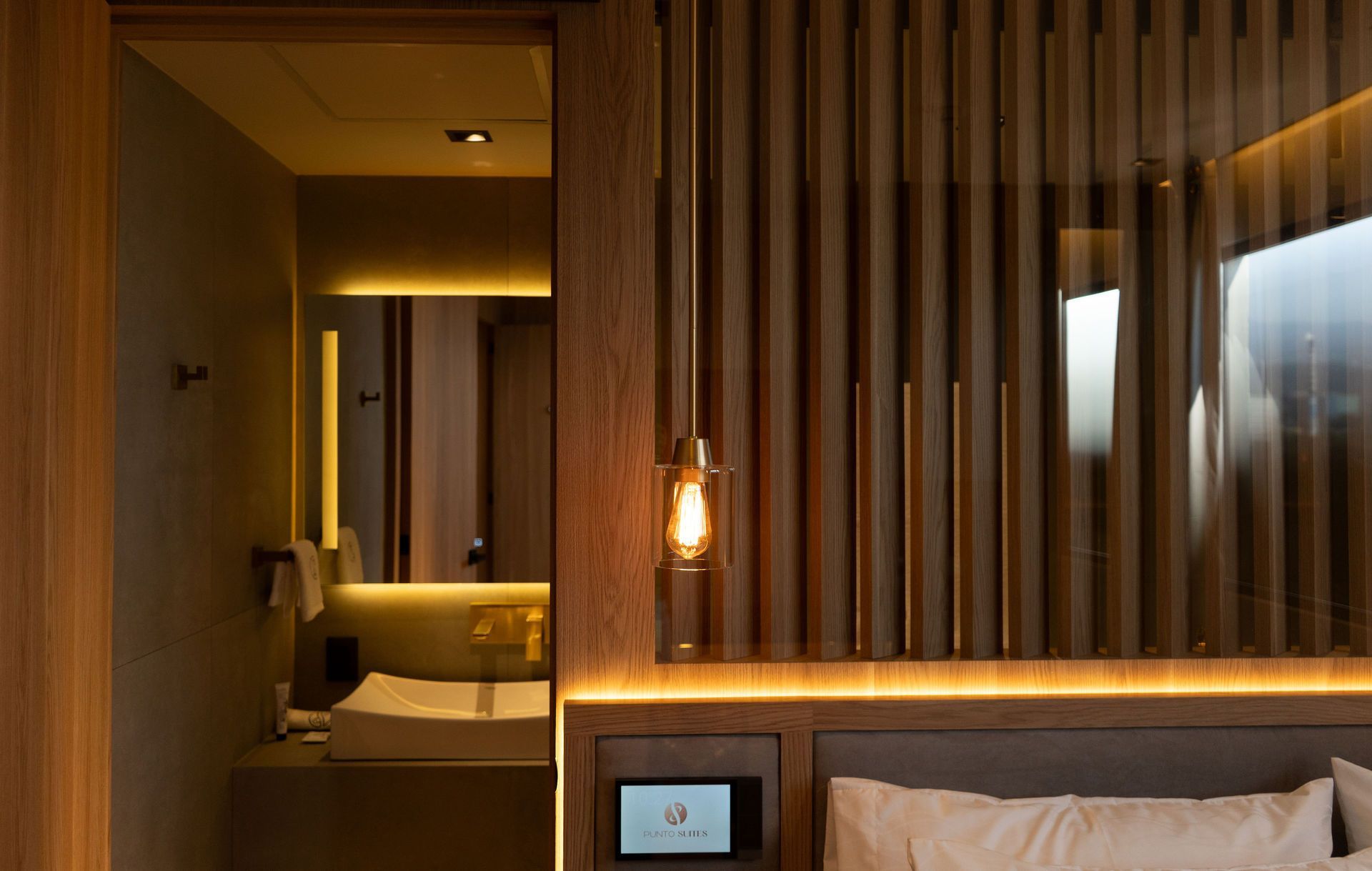 Una habitación de hotel moderna con paredes revestidas de madera, iluminación ambiental cálida, una cama y una vista parcial de un baño contemporáneo.