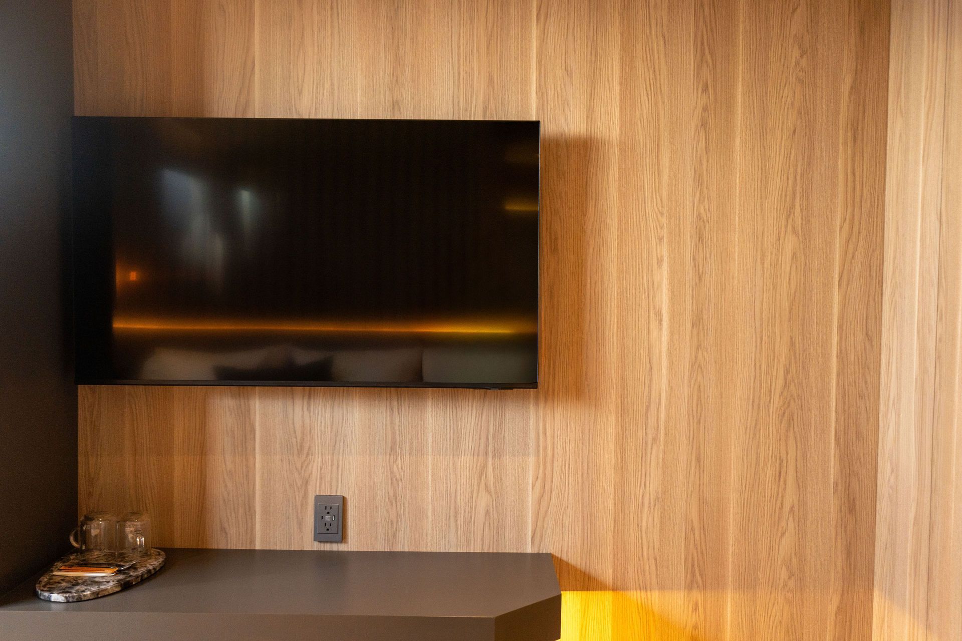 Un televisor de pantalla plana montado en una pared revestida de madera, encima de una estantería oscura con objetos de cristal.