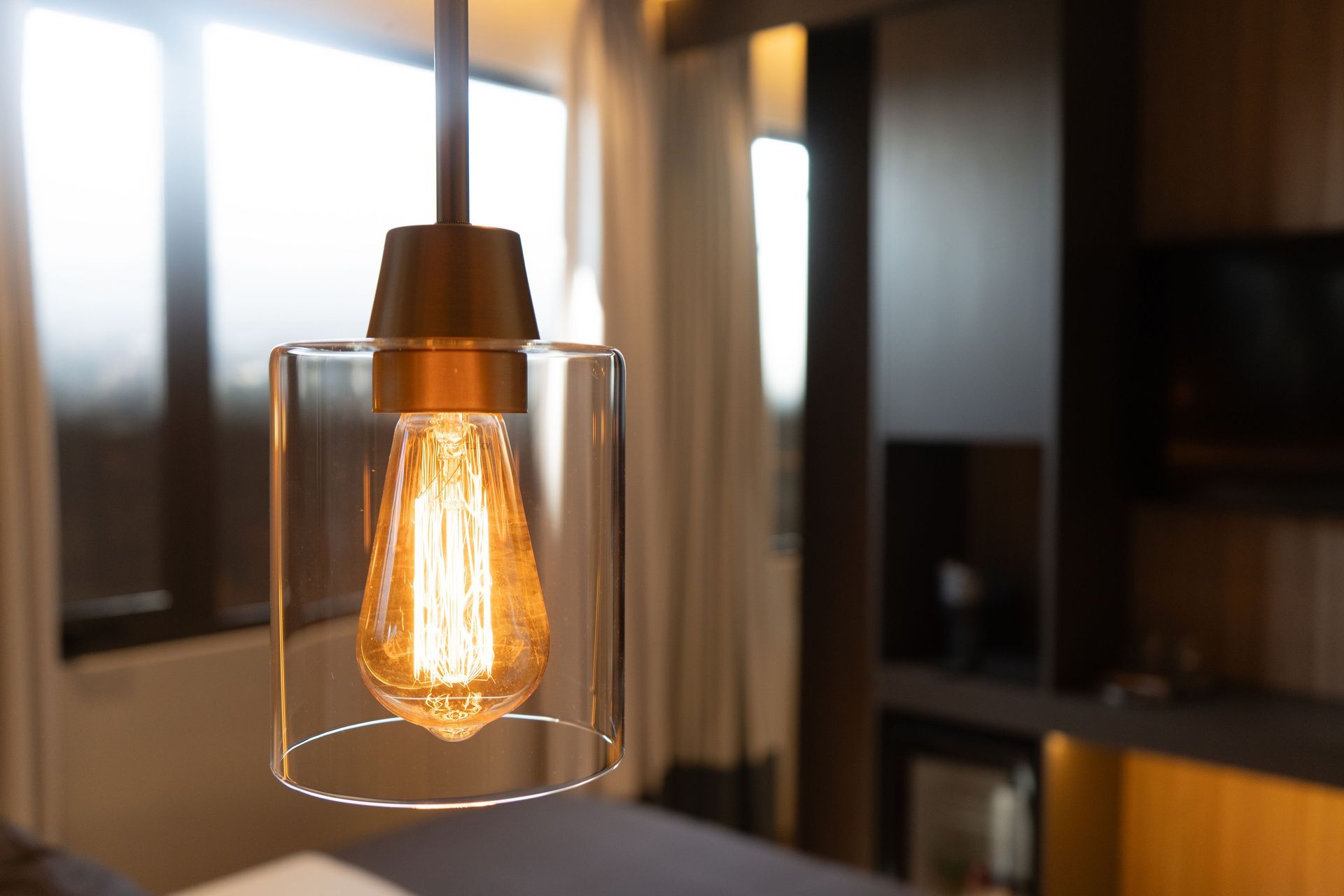 Una bombilla incandescente de estilo Edison cuelga de una pantalla cilíndrica de cristal transparente en una habitación de hotel moderna y con poca luz.