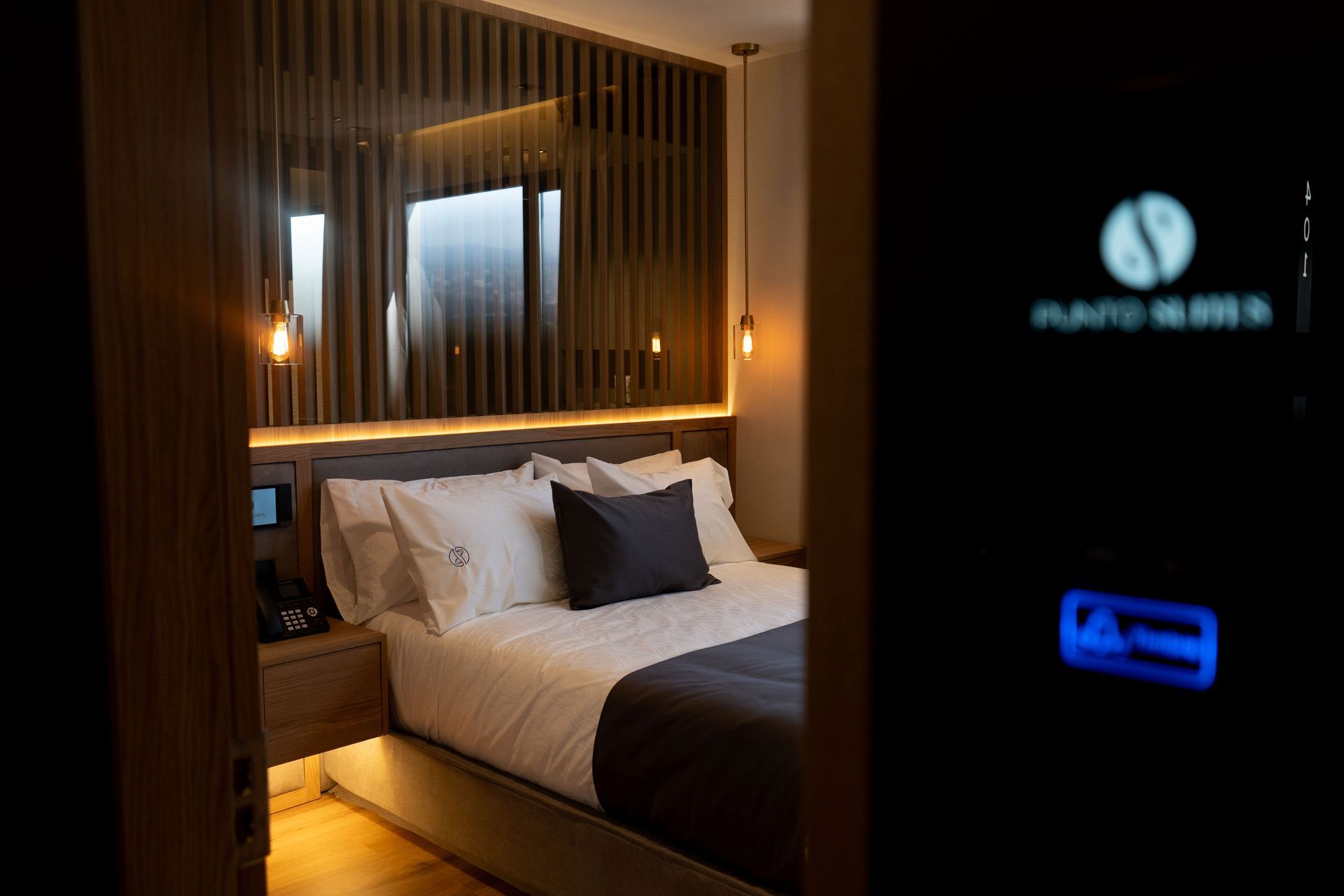 Un dormitorio moderno y con poca luz, con cama, cabecero de madera y lámparas colgantes, visto a través de una puerta oscura.