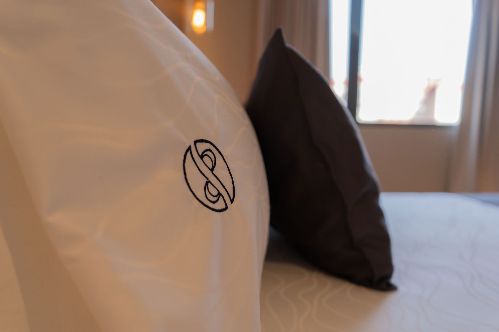 Una almohada blanca con un logotipo circular negro, una almohada decorativa oscura y una ventana al fondo difuminado.