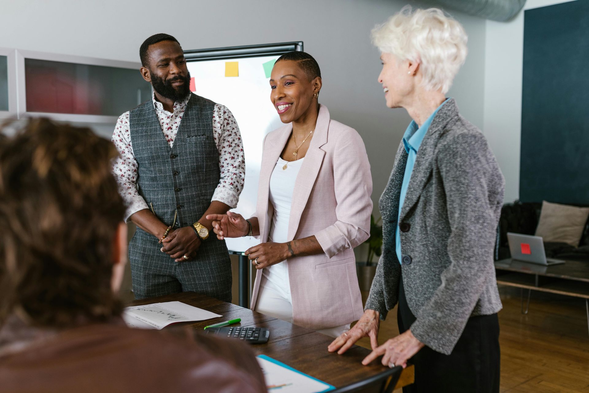 Four people in a meeting, smiling. Man in vest, woman in pink blazer gestures. Whiteboard behind.