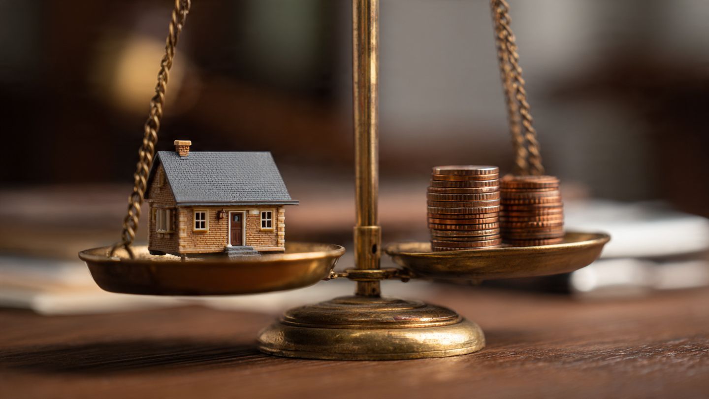 A miniature house sits on one side of a brass balance scale, while a small stack of coins rests on the other side.