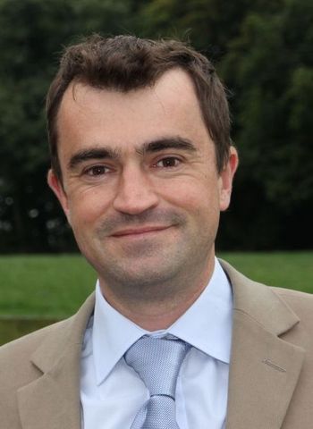 Man in light suit smiles at the camera, light blue tie, outdoors with a green background.