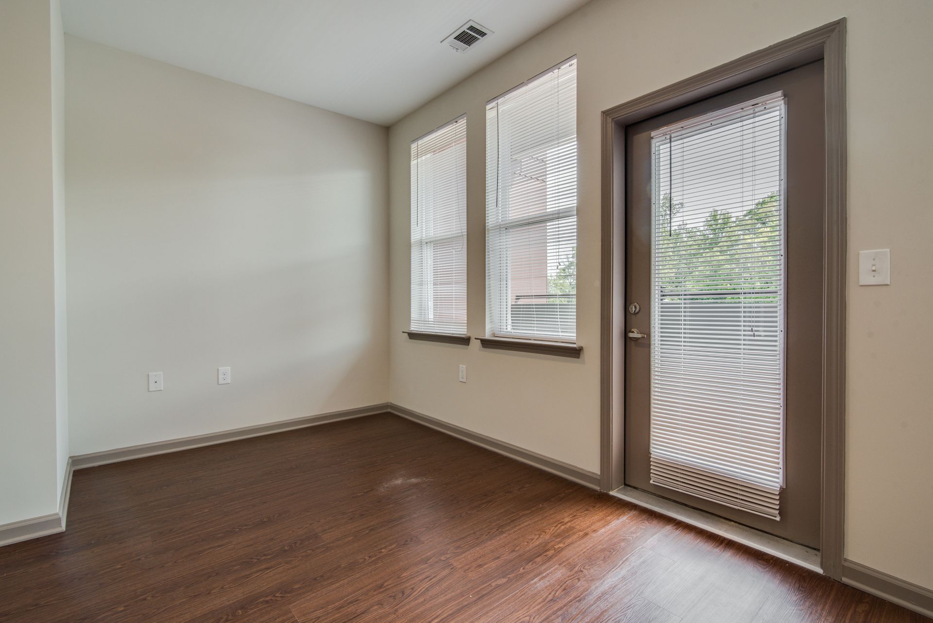 Empty room with dark wood floors, two windows, and a door leading to a balcony.