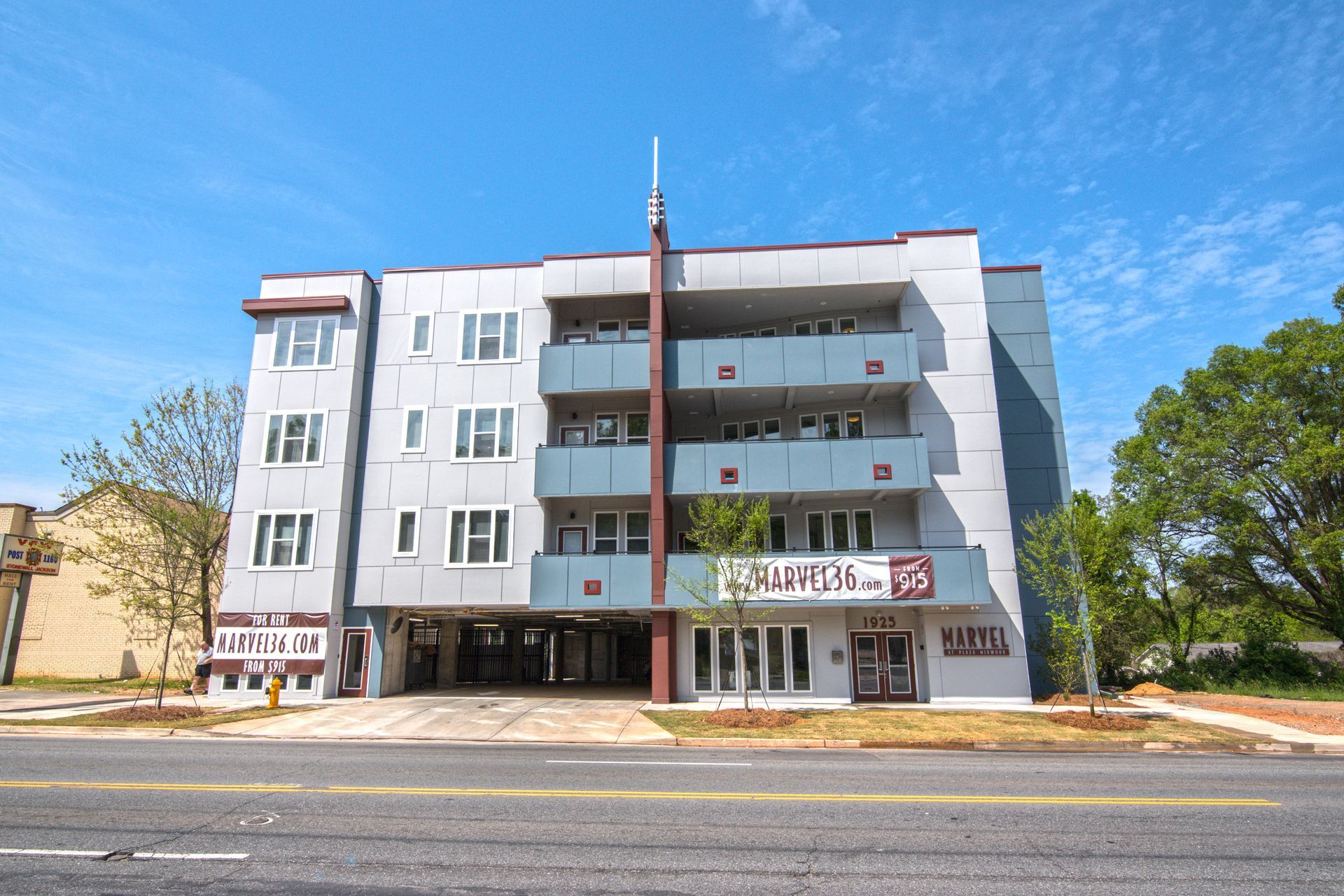 Multi-story apartment building with gray and maroon facade, built over open parking on a sunny day.