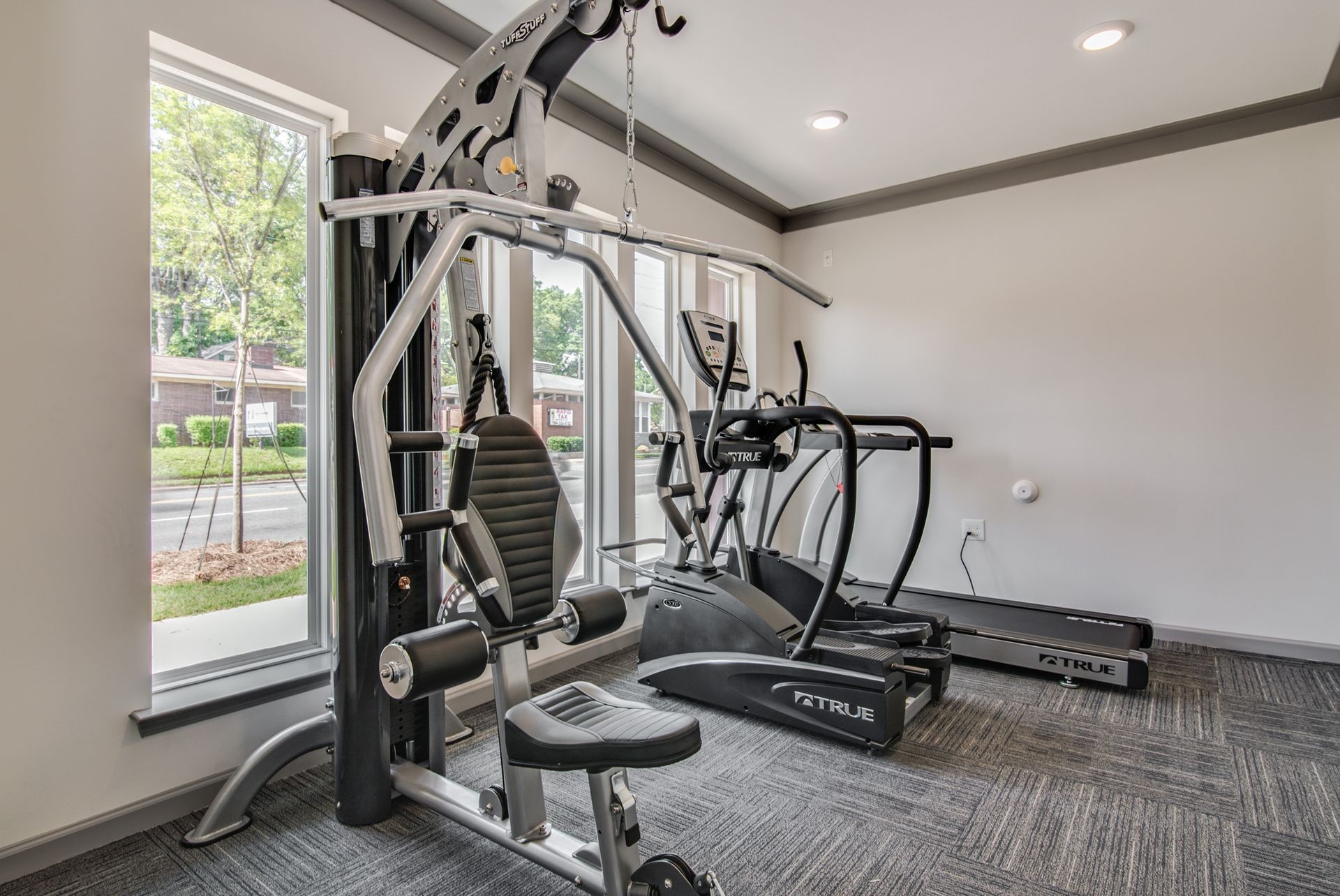 Gym with weight machine, treadmill, elliptical, and window. Gray walls, carpet; fitness equipment.