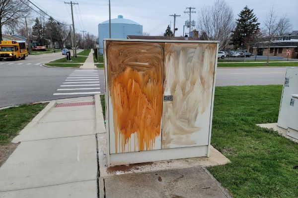 A white box with orange paint on it is on the sidewalk next to a street.