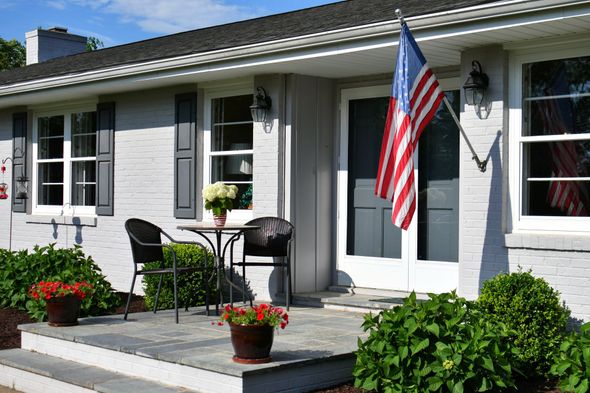 Front porch of a gray house with white trim, potted flowers, patio chairs, and an American flag hanging by the door