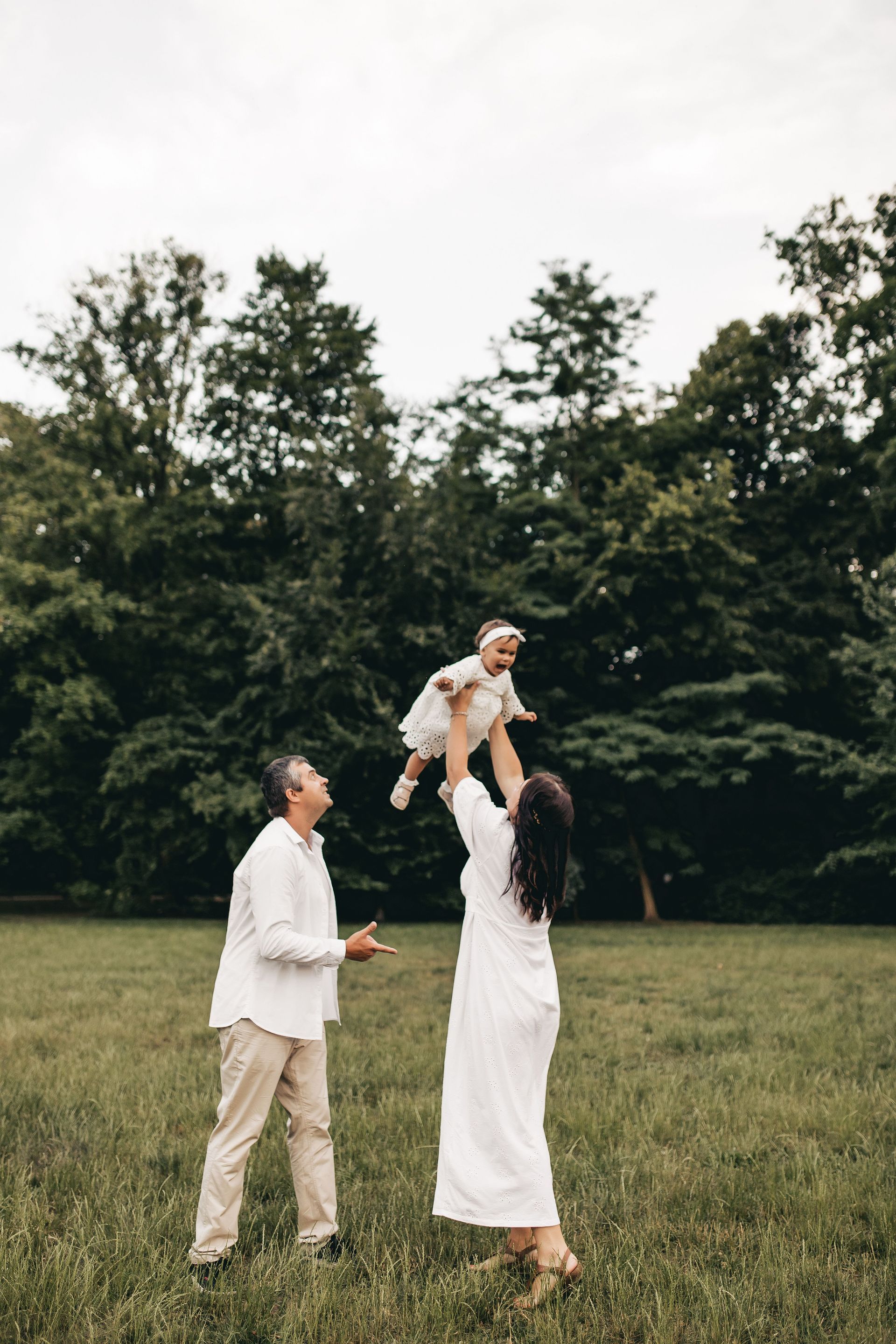 Two adults in white in a grassy field lifting a small child into the air near trees