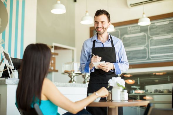 Barista serving a smiling customer at a café counter, with pastries and chalkboard menu in the background