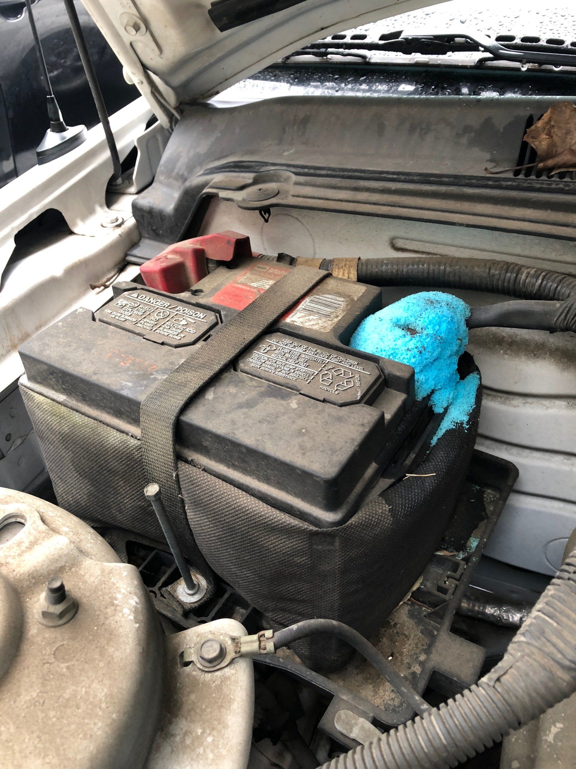 Car battery covered in blue corrosion, held in place by a strap, in engine bay.