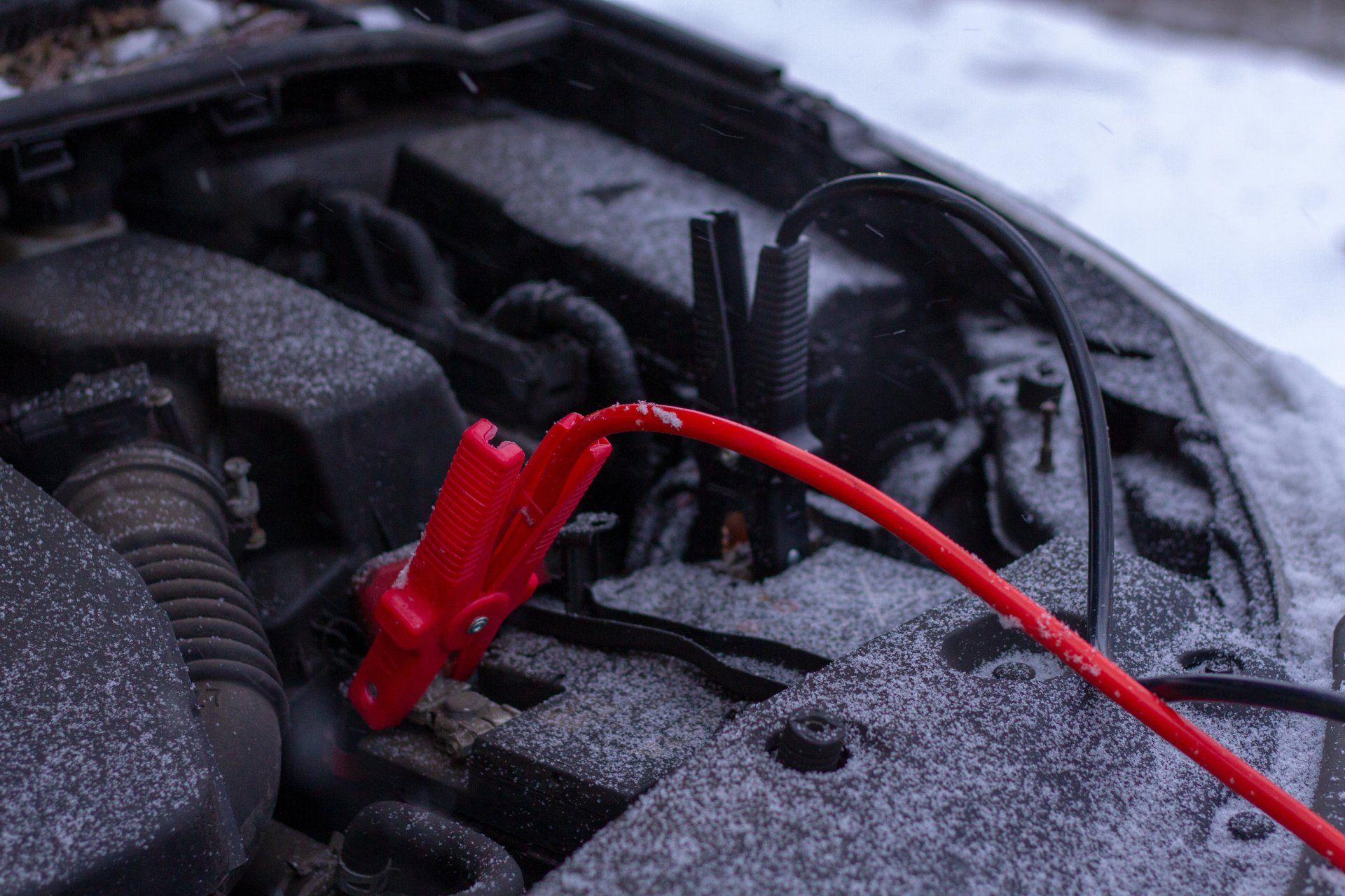 Red jumper cable clamp connected to a car engine in a snowy setting.