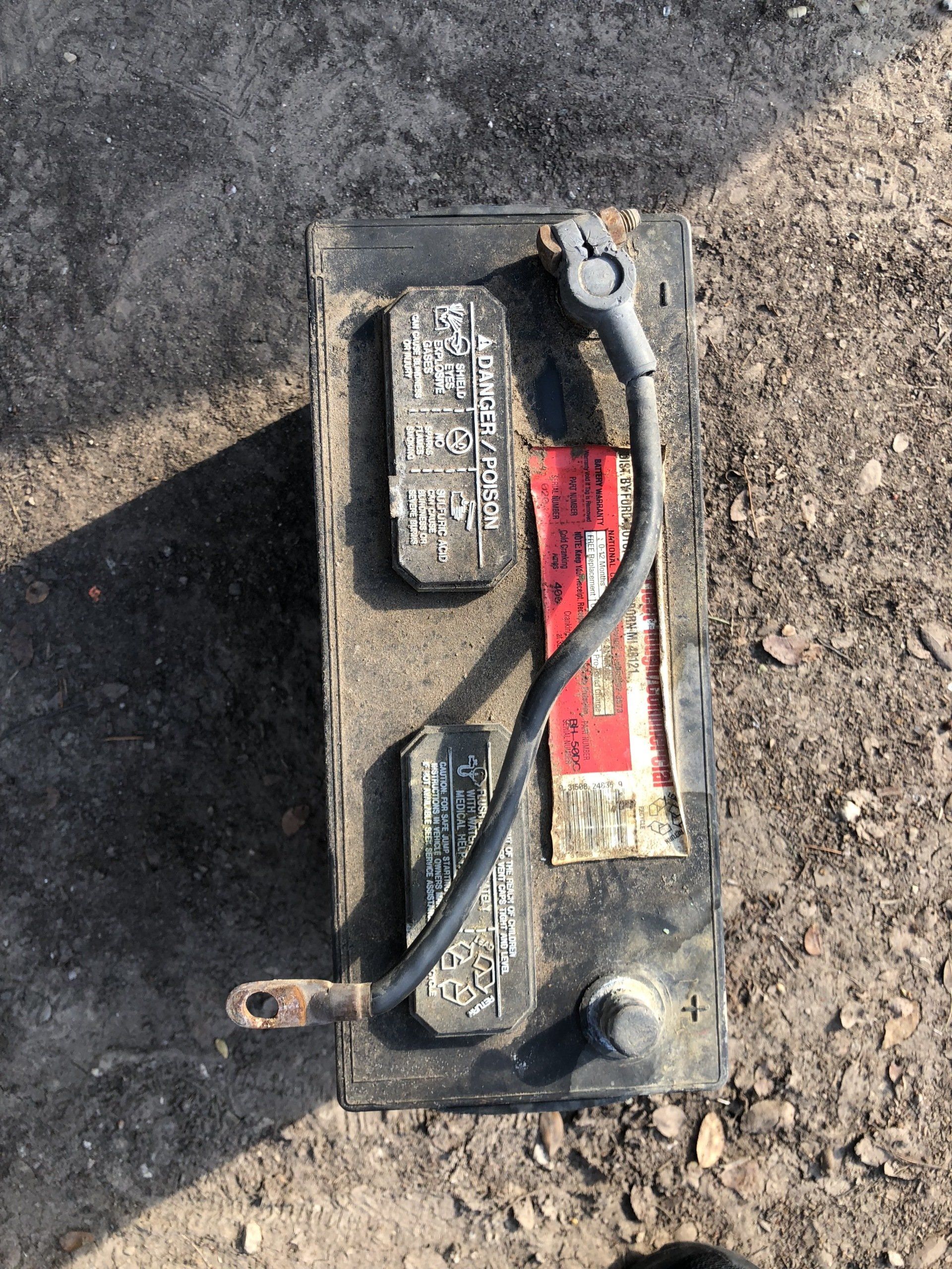 Top-down view of a dirty, rectangular car battery with attached black cables on an outdoor surface.