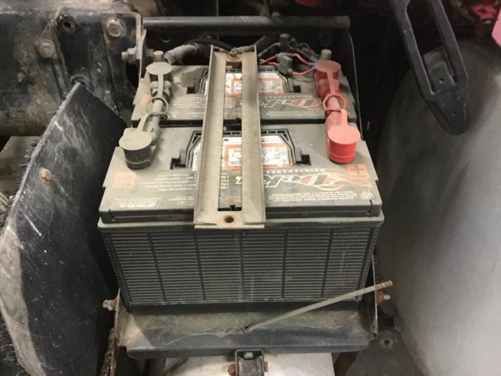 Two rectangular batteries in a vehicle, held by a metal bracket. Red and black terminals are visible.