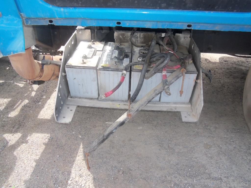 Three large, rectangular batteries in a metal frame under a blue truck. Wires connect the batteries.
