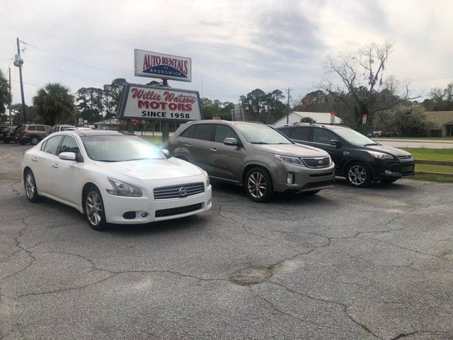 Cars for sale at Willy Wooten Motors. White sedan, gray SUV, and dark sedan parked outside the dealership.