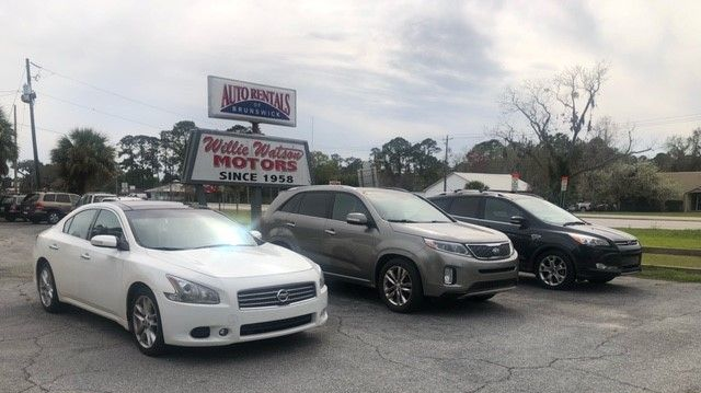 Cars for sale at Willy Wooten Motors. White sedan, gray SUV, and dark sedan parked outside the dealership.