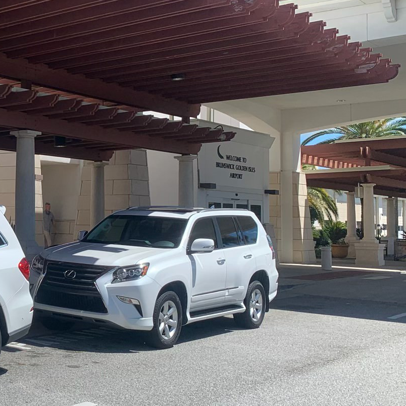 White SUV parked under a brown pergola at a building entrance with pillars.