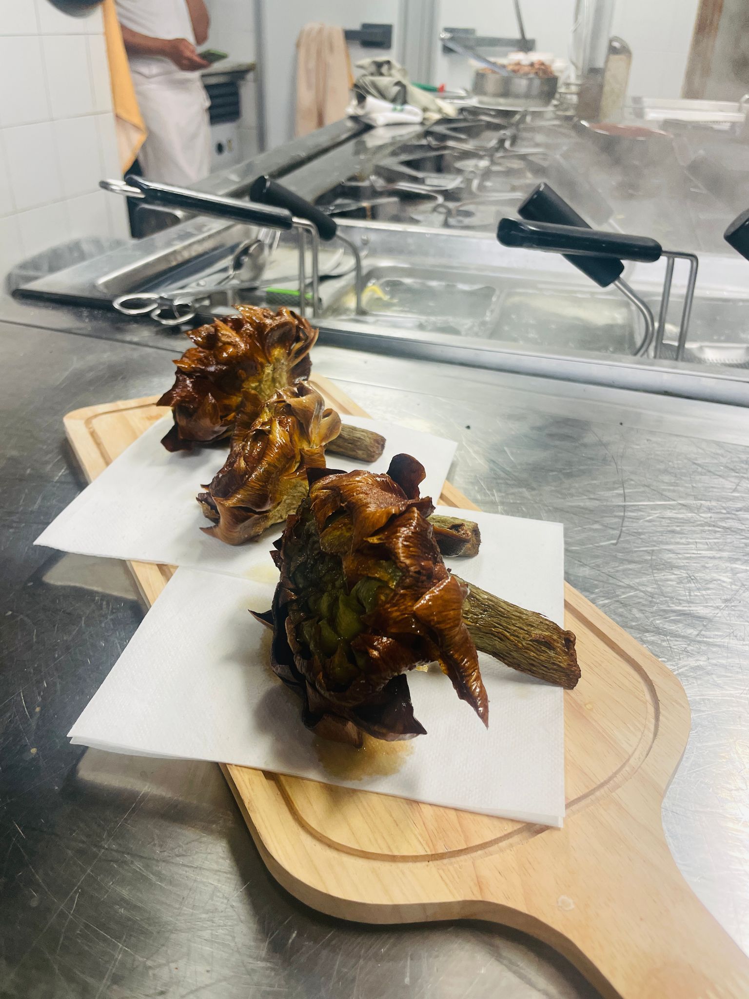 Tre carciofi cotti su carta assorbente, serviti su un tagliere di legno in una cucina commerciale.