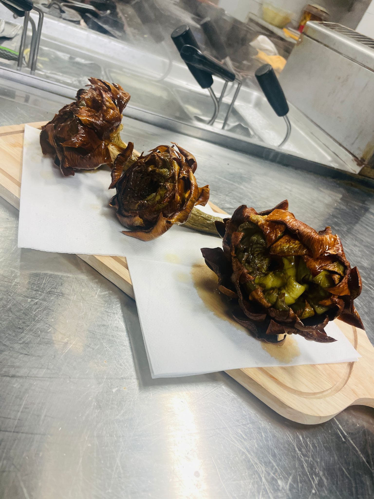 Tre carciofi fritti su carta bianca, appoggiati su un tagliere di legno e appoggiati su una superficie metallica.
