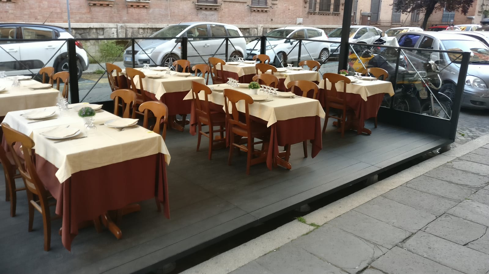 Posti a sedere all'aperto su una piattaforma rialzata, tavoli con tovaglie color crema e gonne marroni, sedie, auto parcheggiate sullo sfondo.
