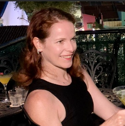 Woman with red hair smiles, sitting at outdoor table with drinks, wearing a black dress.