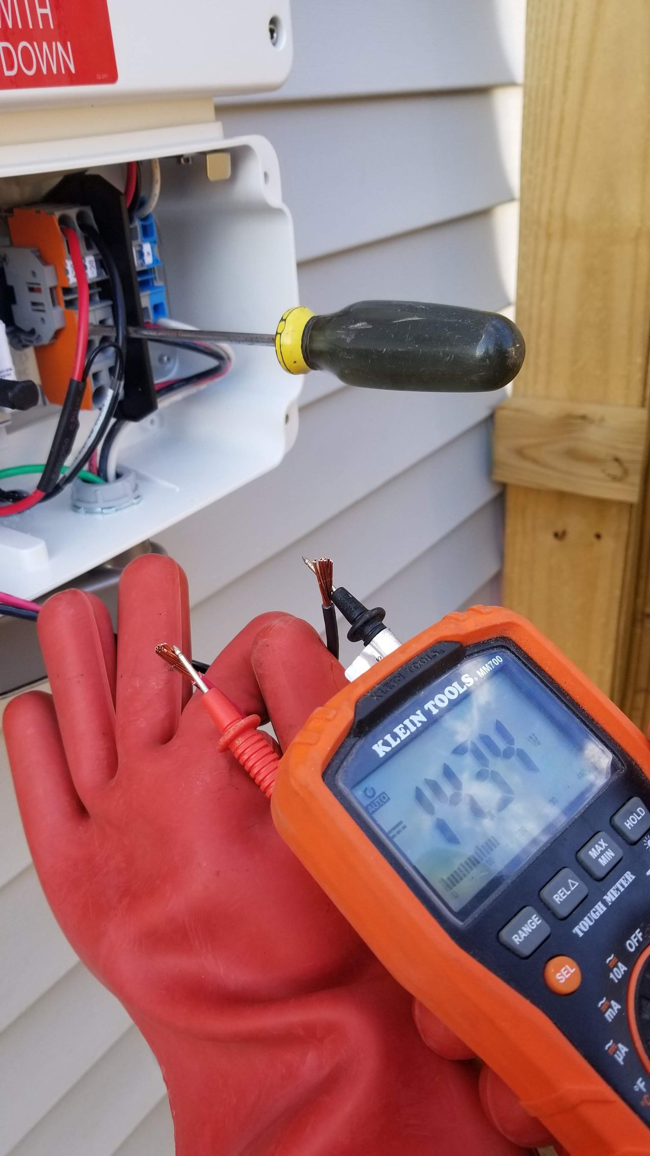A person wearing red gloves is holding a multimeter and a screwdriver