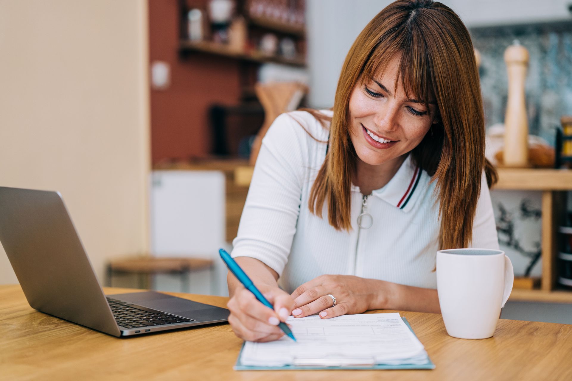 A woman is sitting at a table with a laptop and writing on a clipboard.