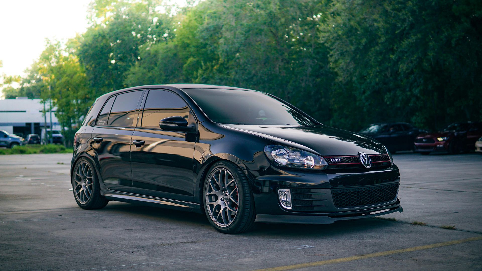 Black Volkswagen Golf GTI parked in a lot, lowered with aftermarket wheels, trees in the background.