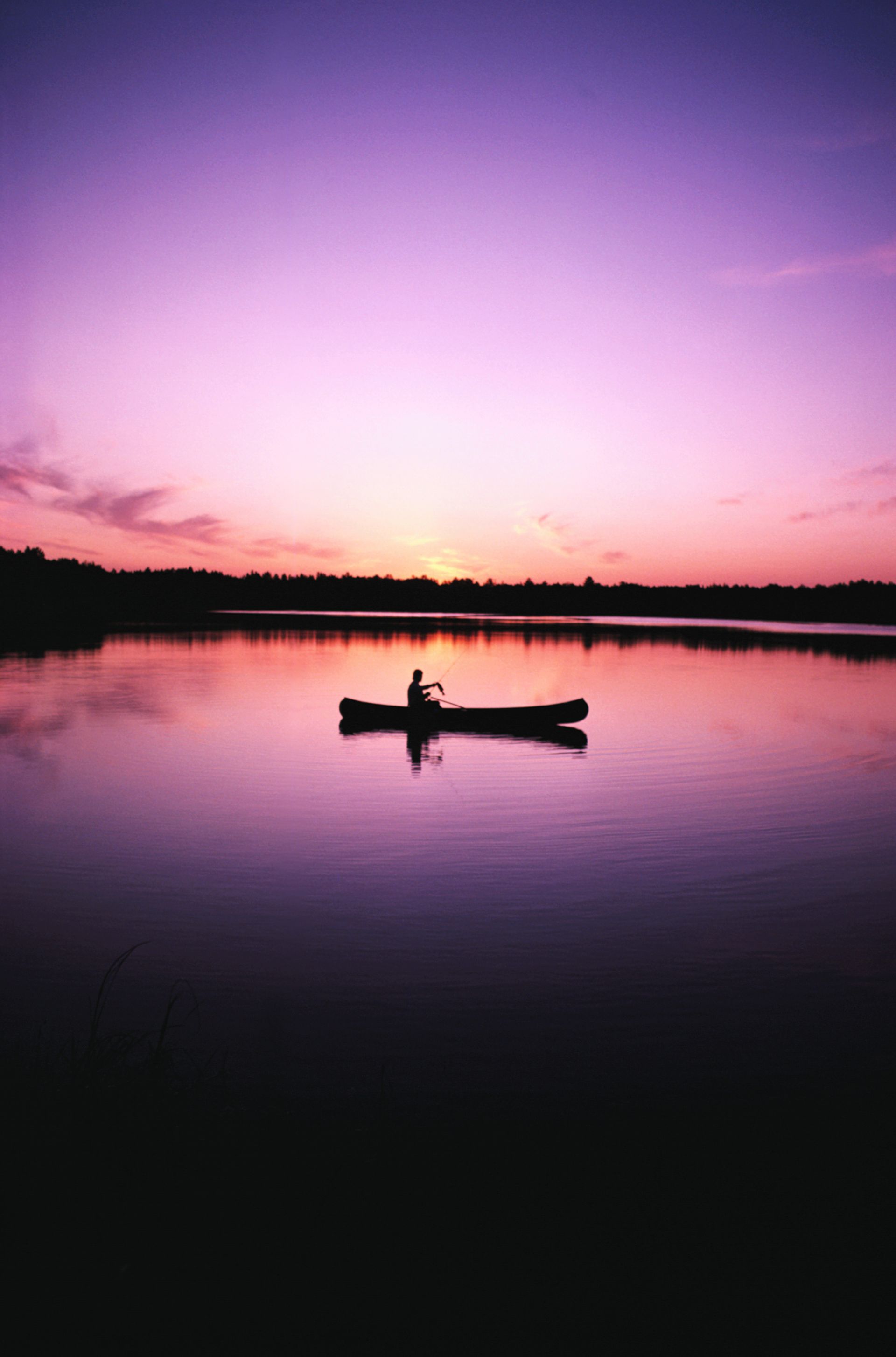 Silhouette of a person canoeing on a calm lake at sunset with vibrant purple and pink hues.