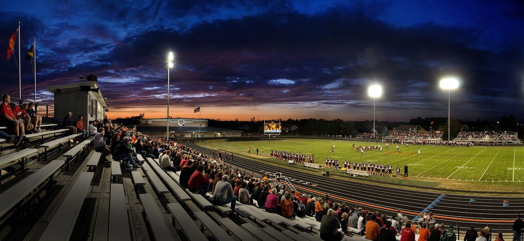Football stadium at dusk; crowd in bleachers, field lit by spotlights, dramatic sky.