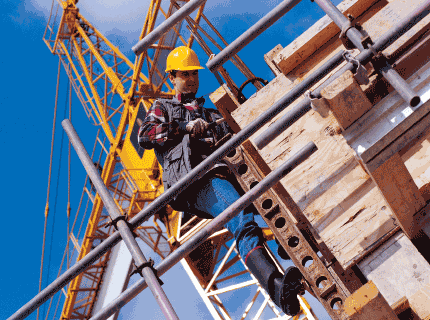 Construction worker in hard hat on scaffolding, blue sky, crane in background.