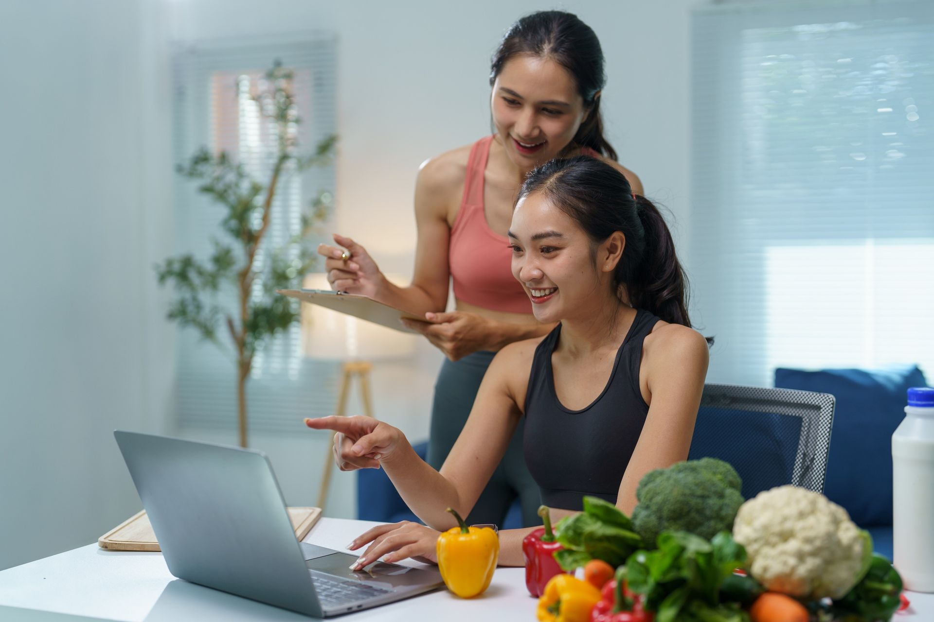 Two women smiling and using a laptop in a bright kitchen with fresh vegetables on the counter