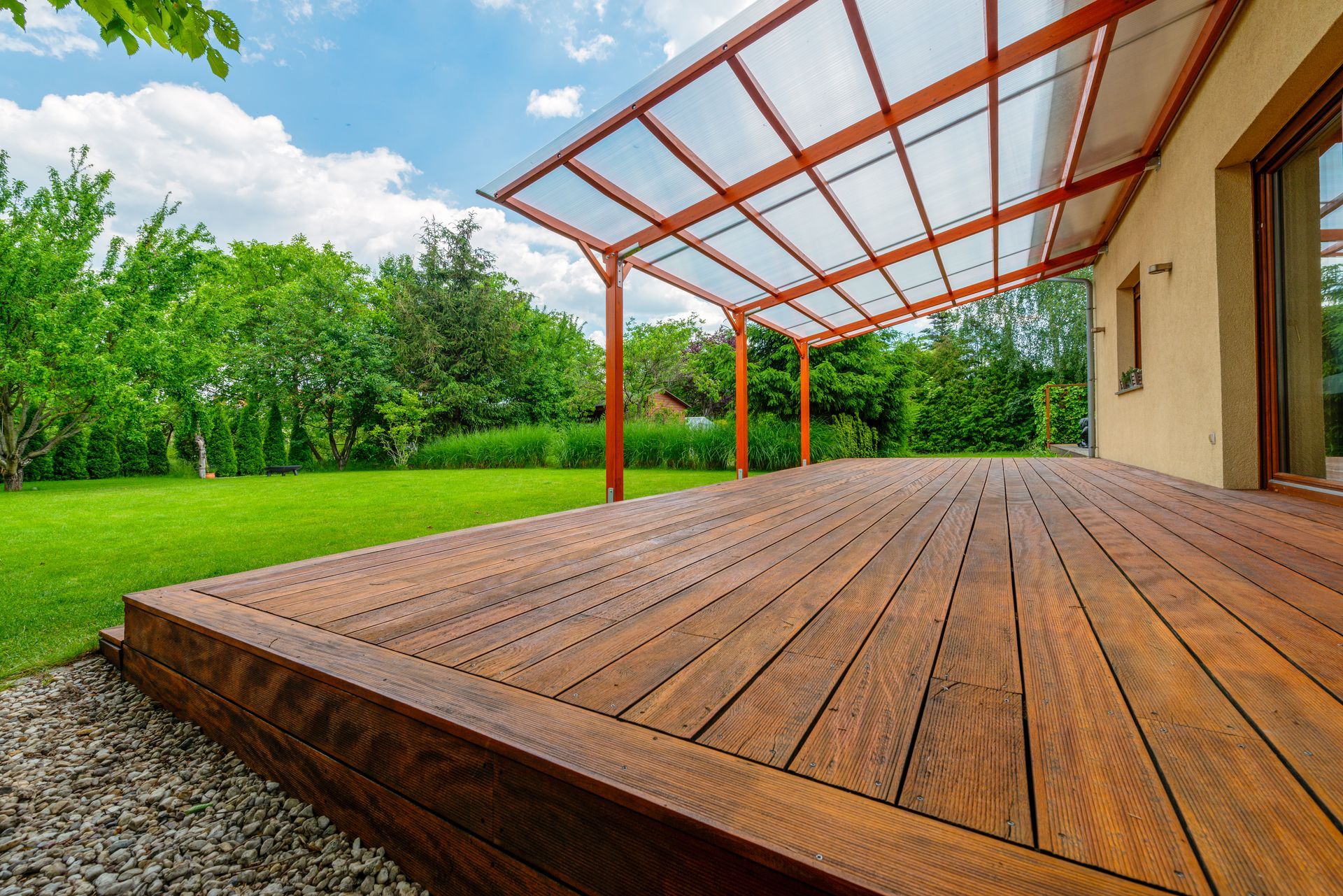 Wooden deck with a glass-roof pergola beside a house, overlooking a green lawn and trees Wooden deck with a glass-roof pergola beside a house, overlooking a green lawn and trees