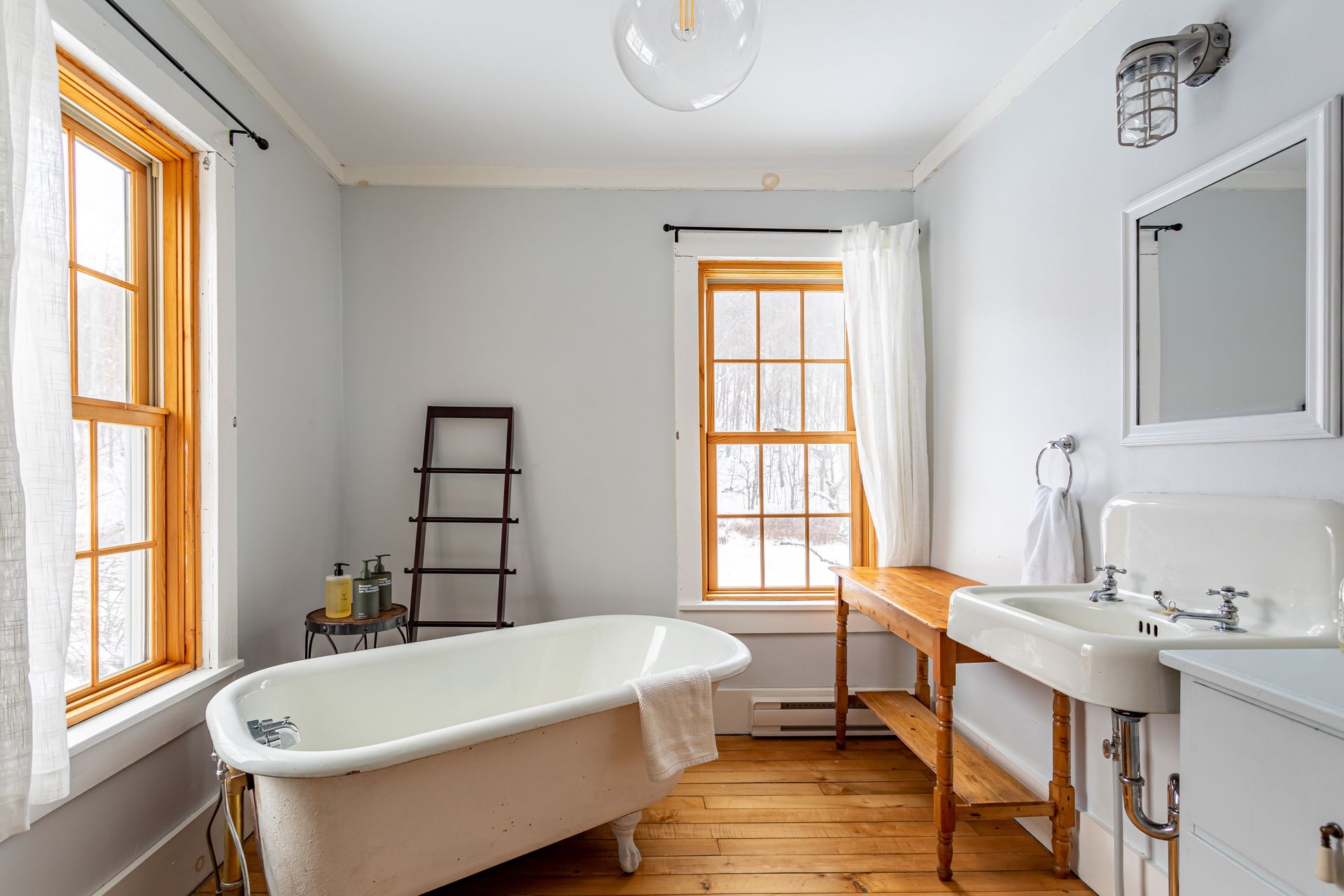 Bright bathroom with freestanding tub, white sink, ladder shelf, and orange-trimmed windows on hardwood floors. Bright bathroom with freestanding tub, white sink, ladder shelf, and orange-trimmed windows on hardwood floors.