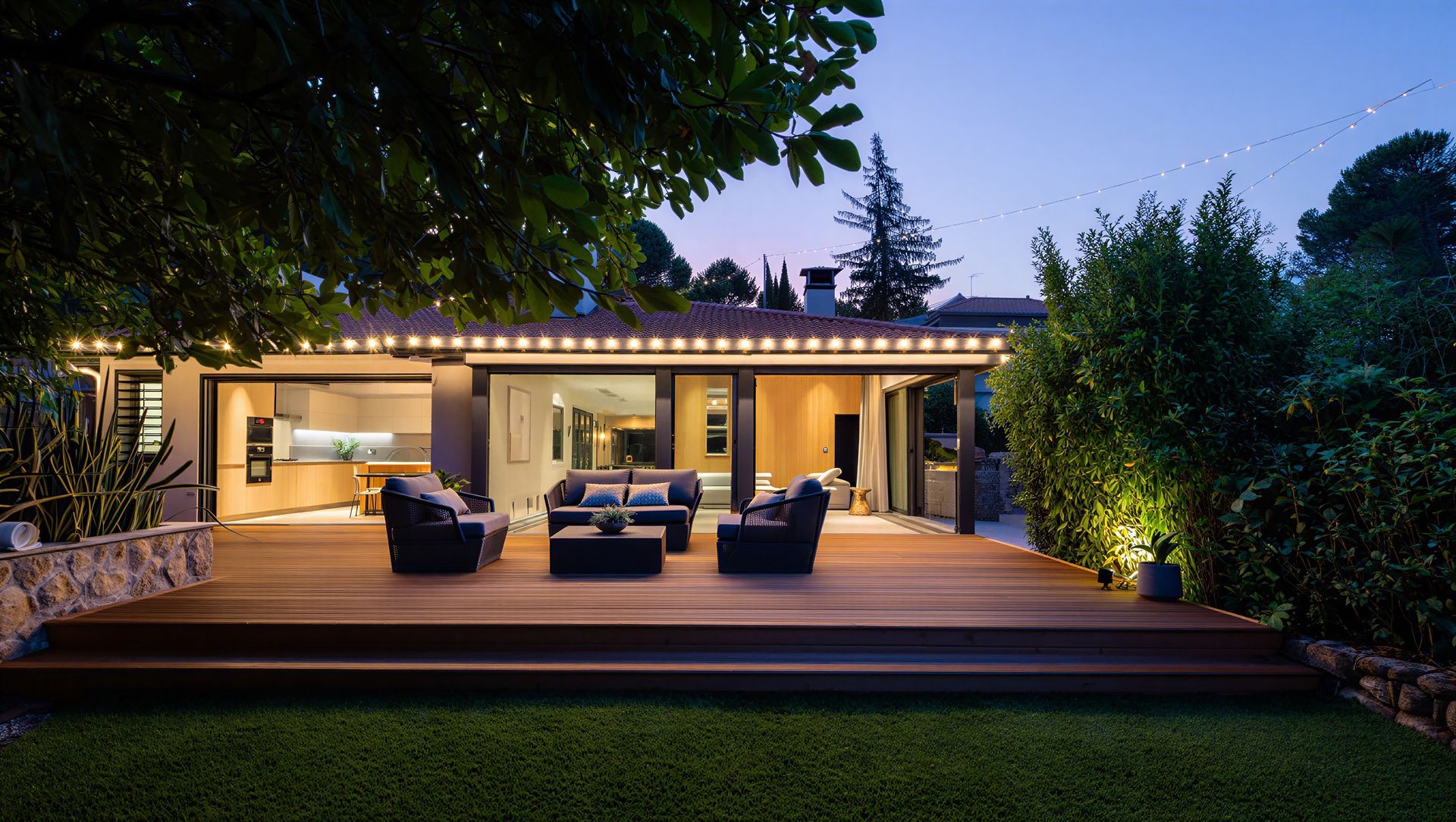 Modern patio with outdoor seating, warm lights, and a wooden deck beside a lit house at dusk Modern patio with outdoor seating, warm lights, and a wooden deck beside a lit house at dusk