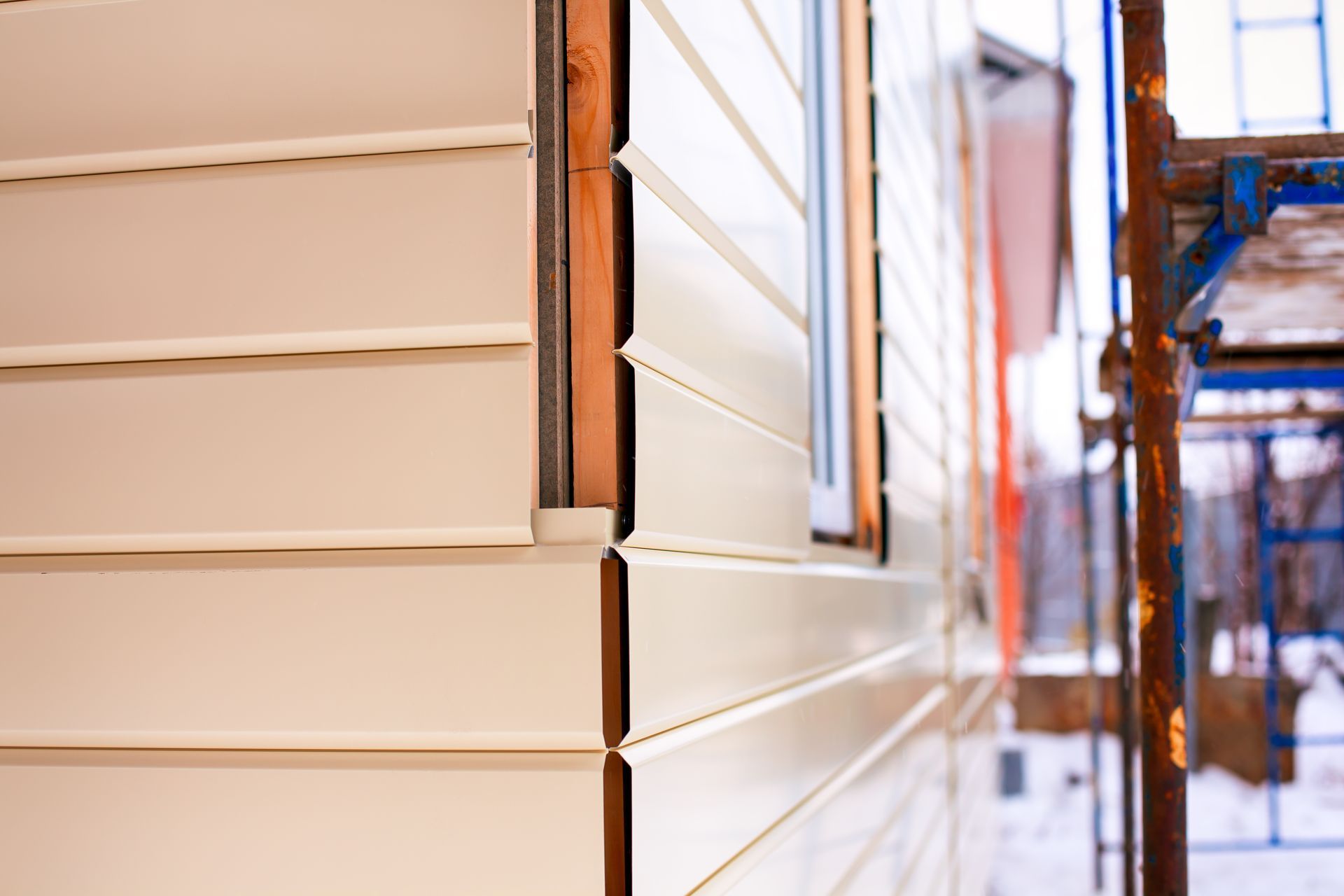 Close-up of beige siding on a building corner with a window, beside a narrow walkway and scaffolding.