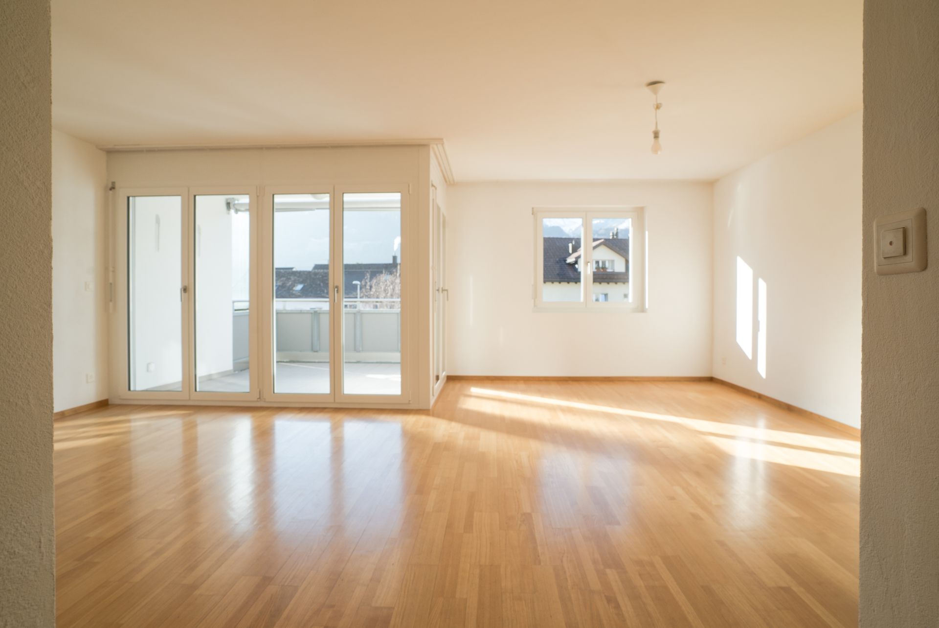 Empty sunlit living room with wooden floors, white walls, and balcony doors/windows. Empty sunlit living room with wooden floors, white walls, and balcony doors/windows.