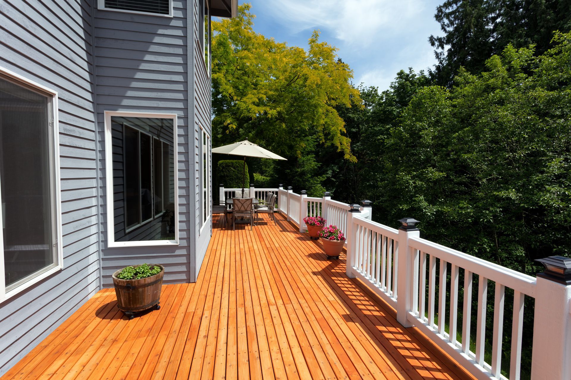 Wooden deck beside gray house, with white railing, potted plants, patio chairs, and trees beyond. Wooden deck beside gray house, with white railing, potted plants, patio chairs, and trees beyond.