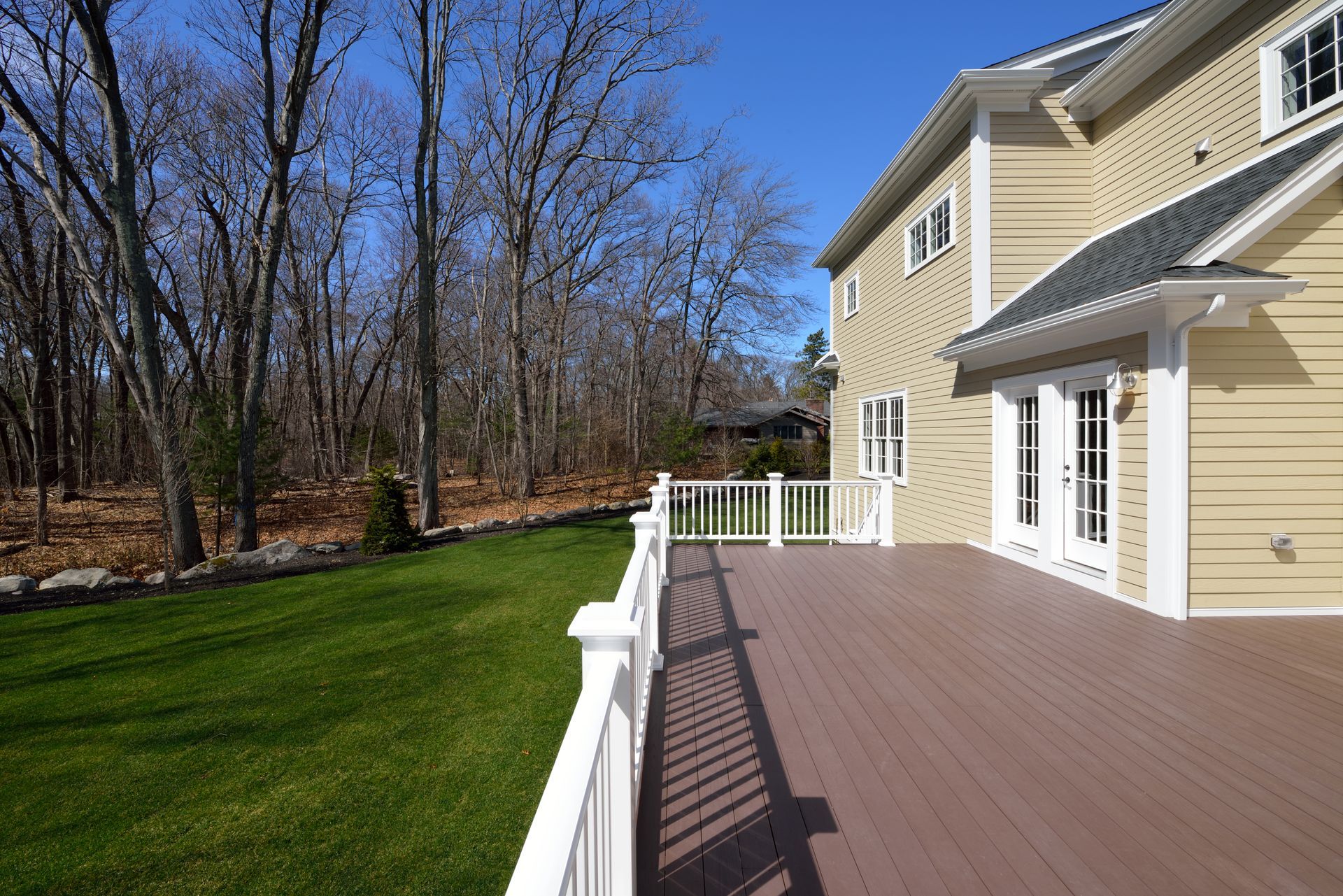 Wide wooden deck beside a beige house, overlooking a grassy yard and leafless trees under a blue sky